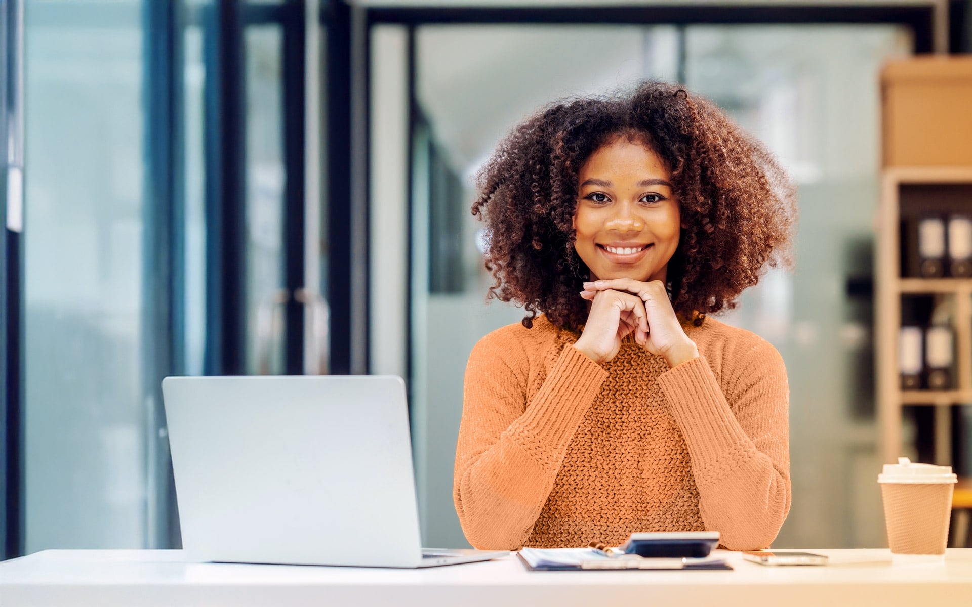Frau im Büro am Schreibtisch die frontal in die Kamera schaut. Auf dem Tisch ein Kaffee und ein Laptop.